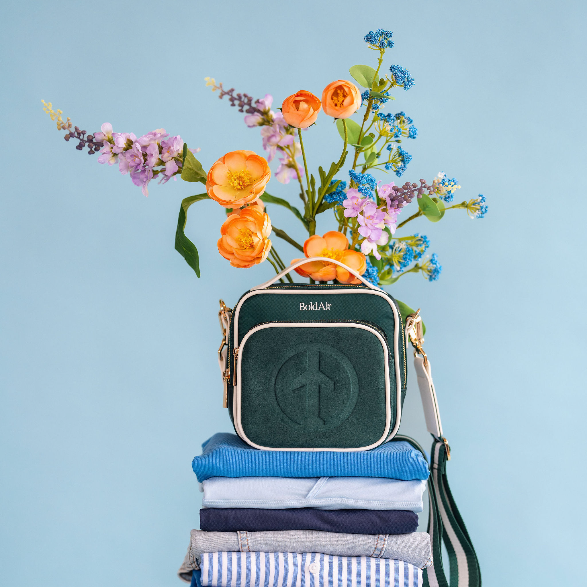 Green BoldAir Boarding Bag with white piping and gold hardware displayed on folded clothing for packing with colorful flowers against a blue background.