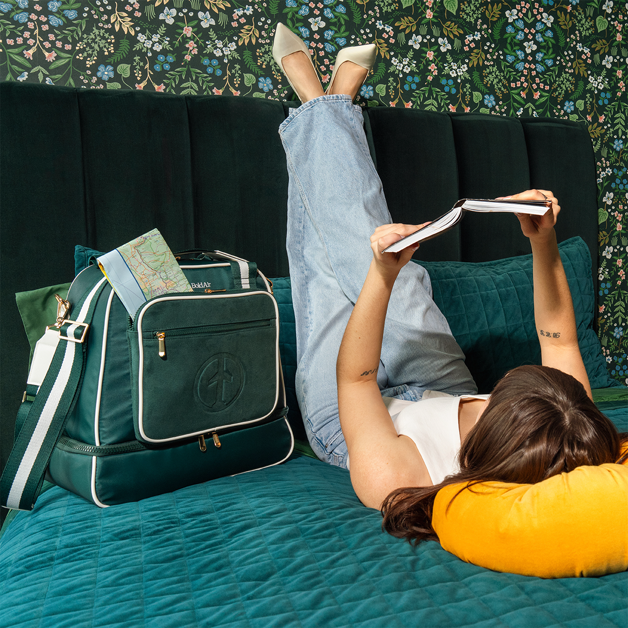 Woman relaxing on a hotel bed with the green BoldAir Layover Bag beside her, a travel map peeking from the women's overnight bag.
