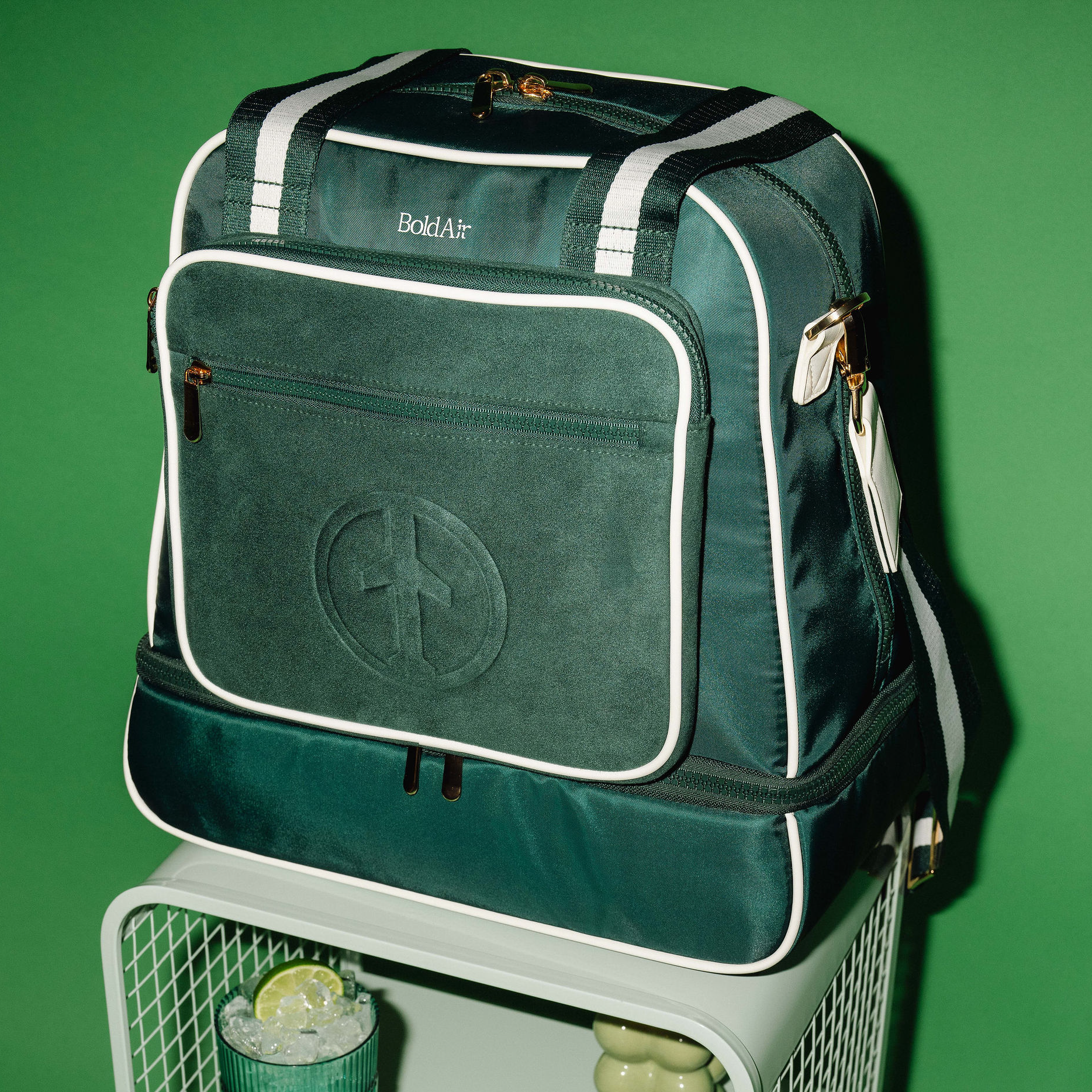 Front view of the green BoldAir Layover Bag with white piping and gold hardware on a hotel nightstand beside an evening cocktail.
