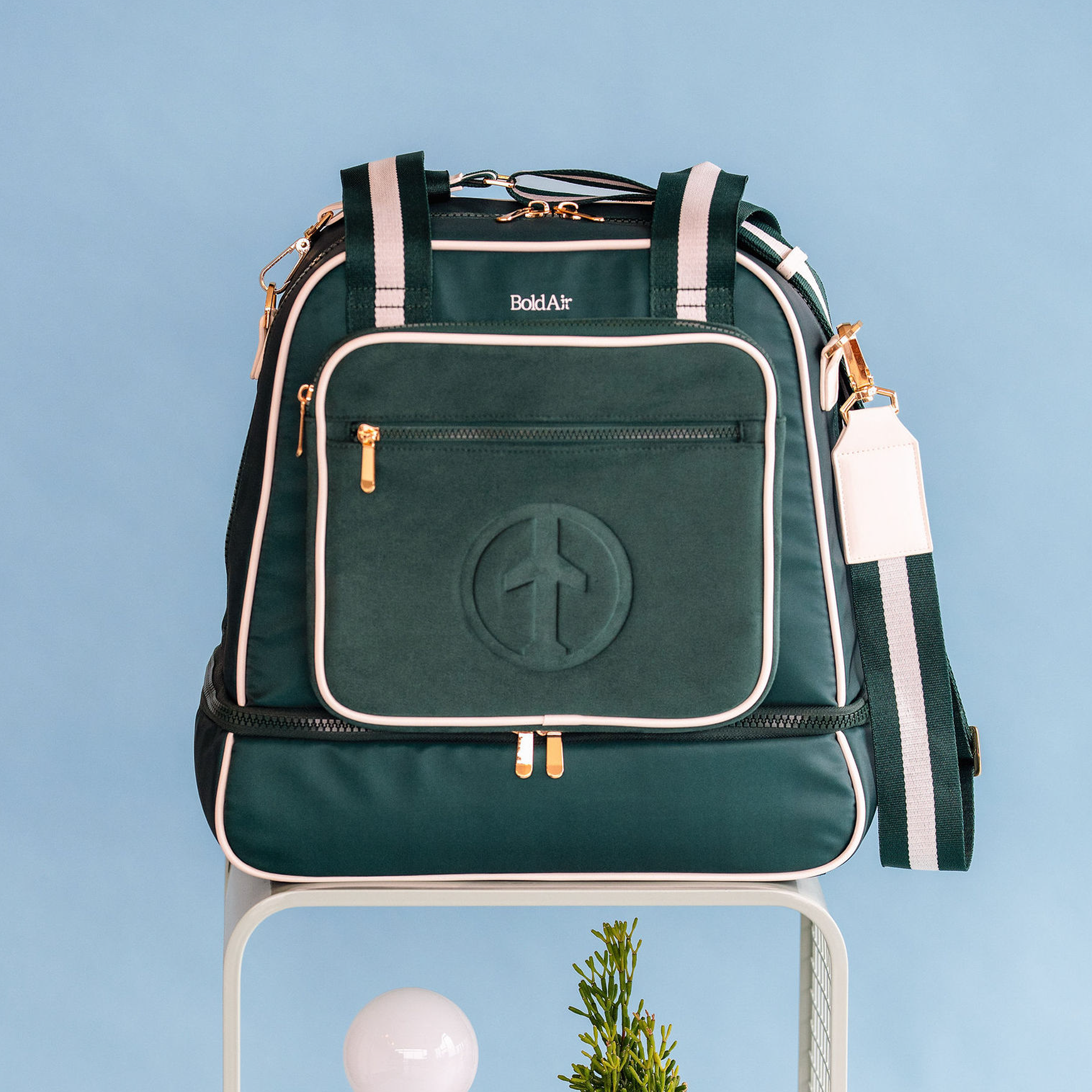 Front view of the green BoldAir Layover Bag with white piping and gold hardware on a hotel nightstand beside travel essentials.