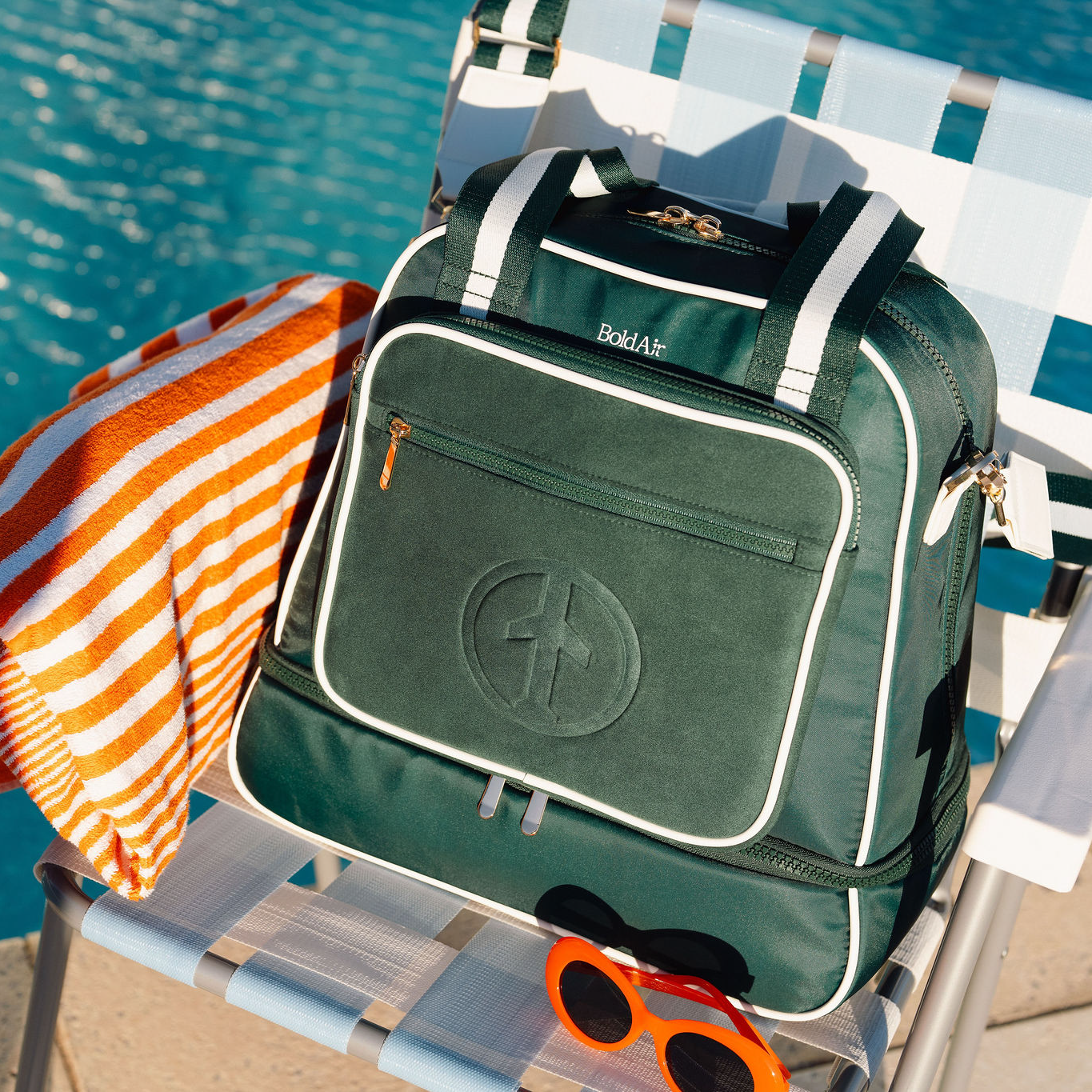 Green BoldAir Layover Bag with white piping and gold hardware on a poolside lounge chair with a beach towel and sunglasses.