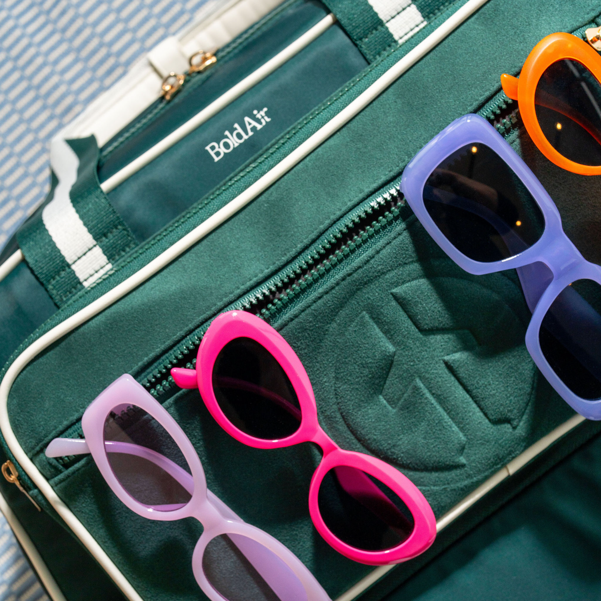 Overhead view of the green BoldAir Stayover Bag with white piping and gold hardware, featuring colorful sunglasses tucked into the exterior pocket.