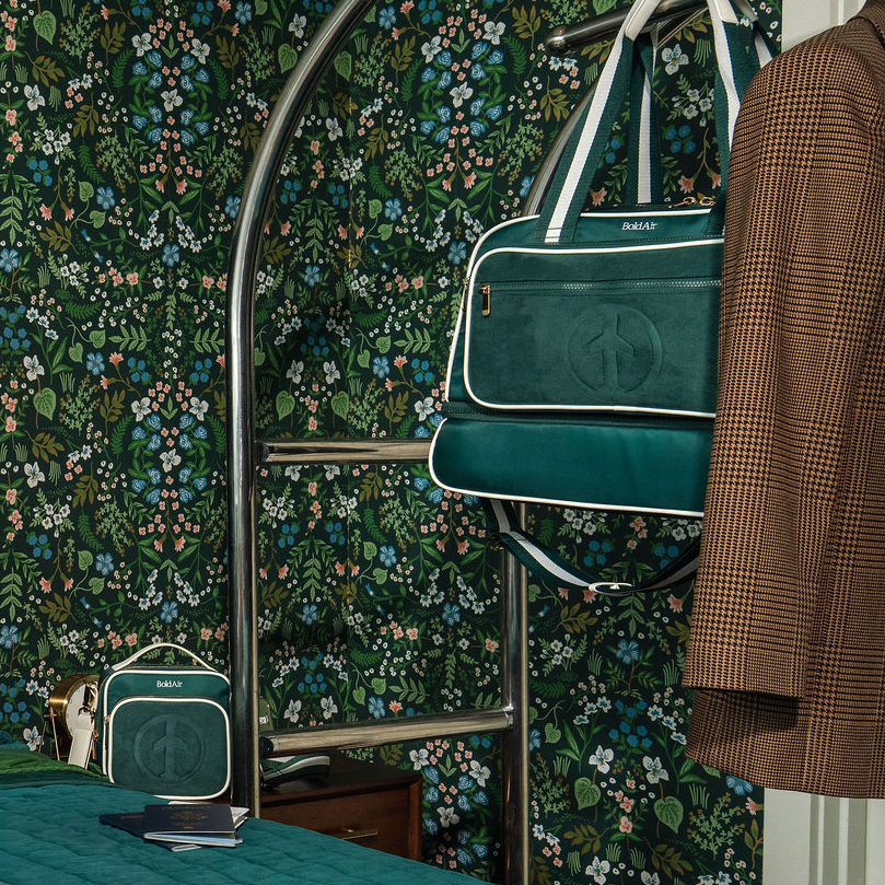 Green BoldAir Stayover Bag hanging from a luggage cart in a boutique hotel room, showcasing the women's weekender bag in a travel setting.