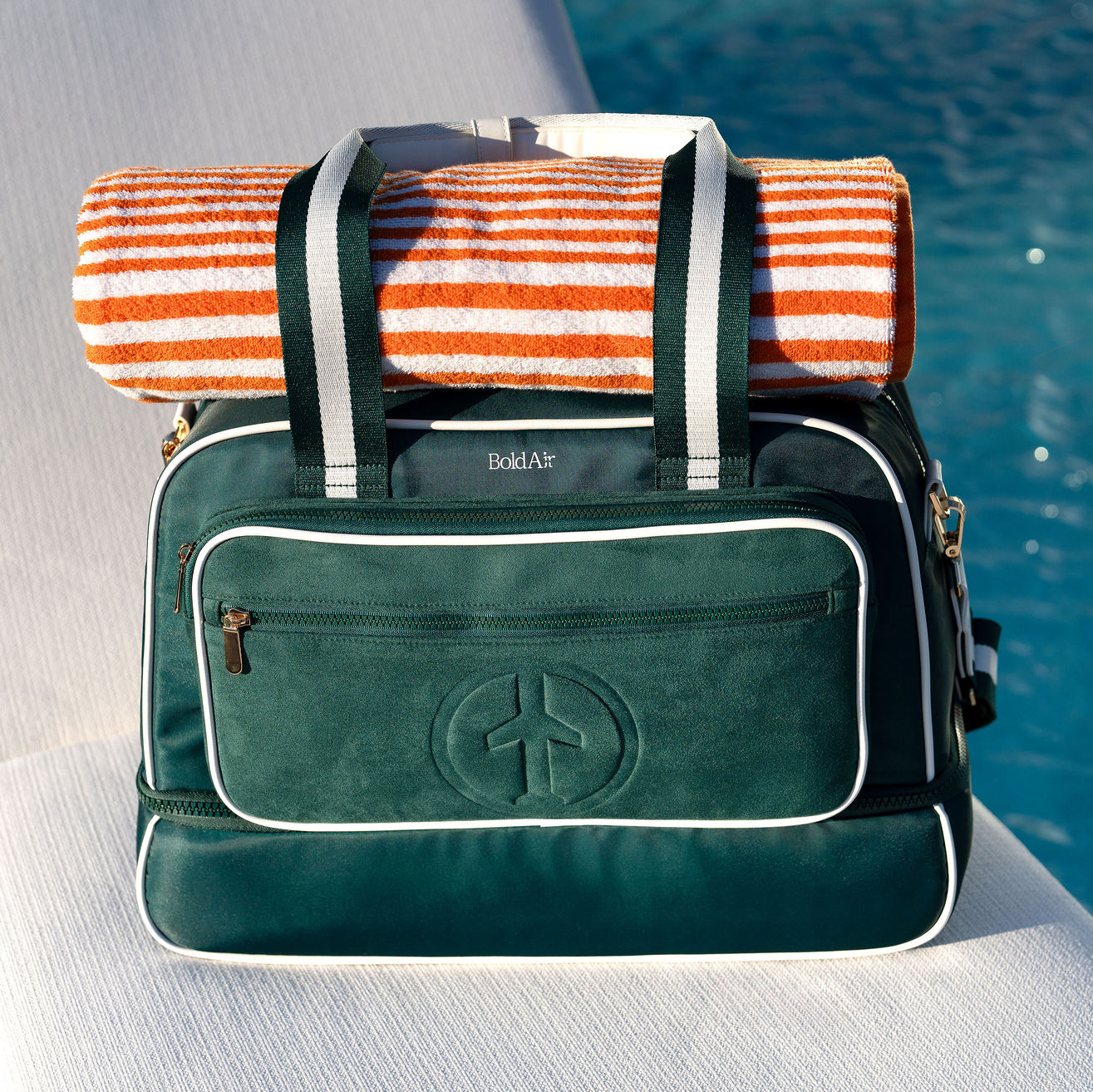 Green BoldAir Stayover Bag with white piping and gold hardware resting on a luxury poolside lounger with a rolled beach towel.